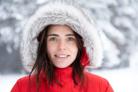 Portrait of a beautiful young smiling brunette with gray eyes in a red jacket with a hood made of faux fur on a winter day against the background of snowy trees.の写真素材