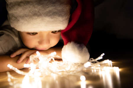 A child in a Santa hat lies on his stomach on the floor in the dark and looks at the Christmas lights. The child looks at the light bulbs and thinks about the gifts. Chin on folded arms.の写真素材