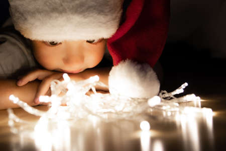 A child in a Santa hat lies on his stomach on the floor in the dark and looks at the Christmas lights. The child looks at the light bulbs and thinks about magic and a fairy tale. Chin on folded arms.の写真素材