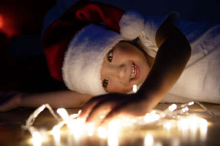 A little boy in a Santa hat and a white T-shirt lies on the floor in the dark and plays with Christmas garlands. The child is laughing, waiting for the New Years holiday with gifts.の写真素材