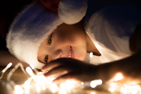 The kid in Santa hat lies on the floor in the dark and plays with Christmas lights. The child...