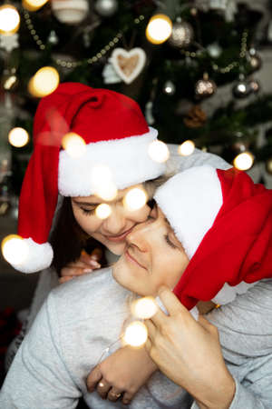 Young loving beautiful couple man and woman in santa hats near christmas tree with lightsの写真素材