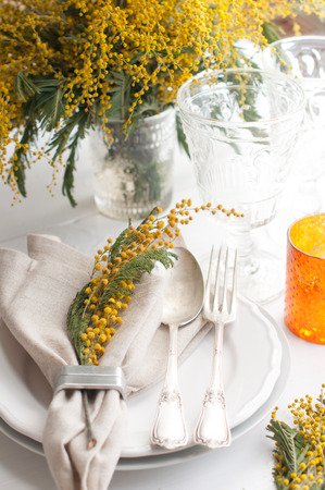 Spring festive dining table setting with yellow mimosa flowers, candles, napkins and vintage cutlery on a white wooden board.の写真素材