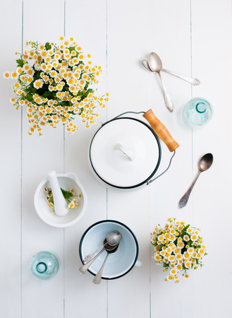 Chamomile flowers, white enamelled cookware, glass bottles, vintage spoons on a white wooden background, cozy home rustic decor, vintage country livingの写真素材