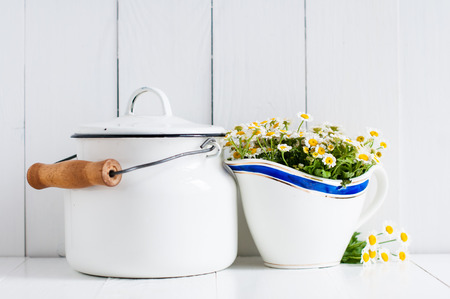 Chamomile flowers in vintage ceramic gravy boat and enamel milk can on white wooden background, home...