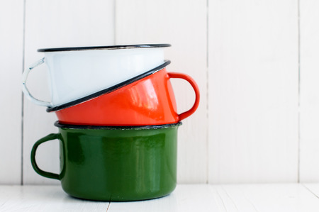 A stack of three bright colorful enameled mugs on white painted wooden table, kitchenware and decor, rustic vintage kitchen backgroundの写真素材