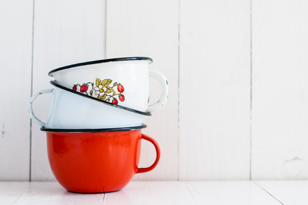 A stack of three bright colorful enameled mugs on white painted wooden table, kitchenware and decor,...