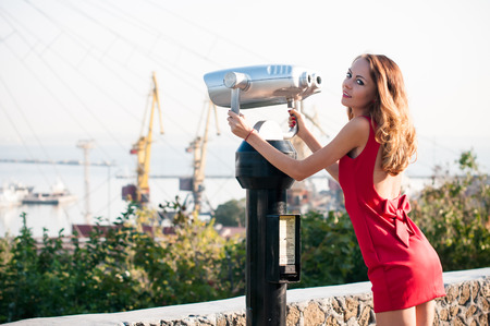 Beautiful young slim woman in a morning light on a city street, lady in red dress has fun watching at the tower viewer to the port panoramaの写真素材