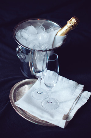 A bottle of chilled champagne in an ice bucket and two glasses on a bed, dark tonesの写真素材