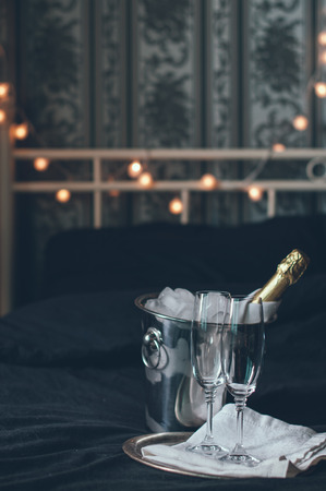 A bottle of chilled champagne in an ice bucket and two glasses on a bed, dark tonesの写真素材