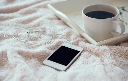 White smart phone with headphones and a cup of coffee on a tray on the bedの写真素材