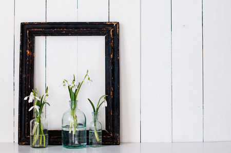 White spring flowers snowdrops in vintage glass bottles and a blck frame on an white barn wall backgroundの写真素材