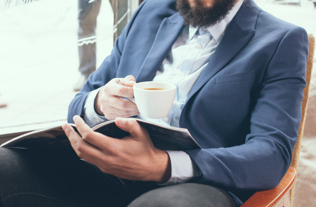 Young businessman in a blue jacket with a cup of coffee, reading a magazine in a cafeの写真素材