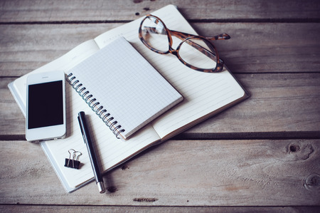 Hipster home office tabletop: papers and notebooks, reading glasses, smart phone, pen on an old wooden board backgroundの写真素材