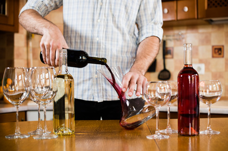 Man pouring red wine from bottle into a decanter, home kitchen interior, bottles of wine