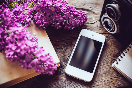 Books, notebooks, vintage camera, white smart phone and lilac flowers on an old wooden board background, hipster lifestyle arrangementの写真素材