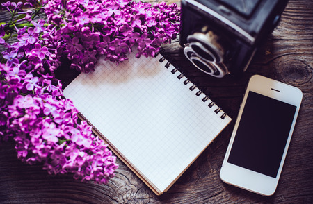 Books, notebooks, vintage camera, white smart phone and lilac flowers on an old wooden board background, hipster lifestyle arrangementの写真素材