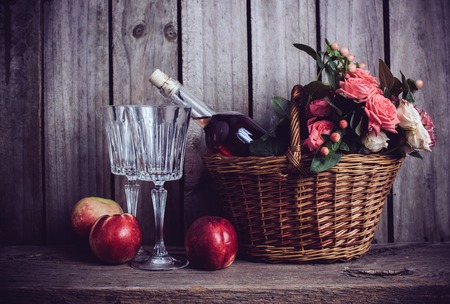 Rustic still life, fresh natural pink roses in a wicker basket  and a bottle of rose wine with two wineglasses and nectarines on an old wooden barn board background. Flowers and fruits for vintage wedding.