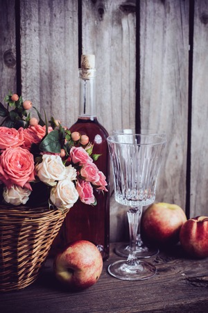 Rustic still life, fresh natural pink roses in a wicker basket  and a bottle of rose wine with two wineglasses and nectarines on an old wooden barn board background. Flowers and fruits for vintage wedding with copy space.の写真素材