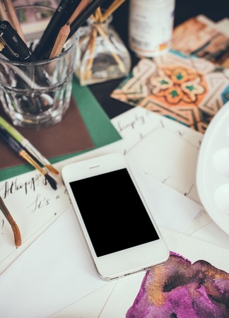 Smartphone on the table in the artist's studio, watercolor paints, brushes and sketches, palette and painting tools. Arts and modern technology, hipster style.の写真素材