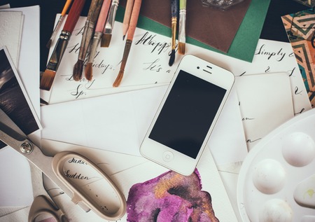 Smartphone on the table in the artist's studio, watercolor paints, brushes and sketches, palette and painting tools. Arts and modern technology, hipster style.の写真素材