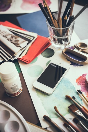 Smartphone on the table in the artist's studio, watercolor paints, brushes and sketches, palette and painting tools. Arts and modern technology, hipster style.の写真素材