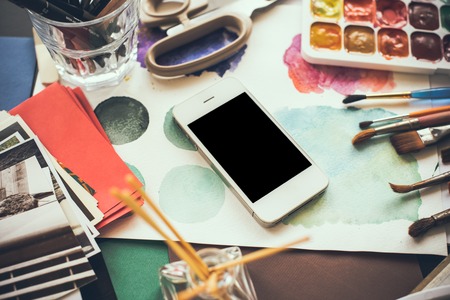 Smartphone on the table in the artist's studio, watercolor paints, brushes and sketches, palette and painting tools. Arts and modern technology, hipster style.の写真素材