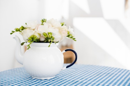 Lovely fresh bouquet of white summer roses and freesias in vintage enamel tea pot on a table with blue vichy tablecloth. Retro style interior decoration with copy space.の写真素材