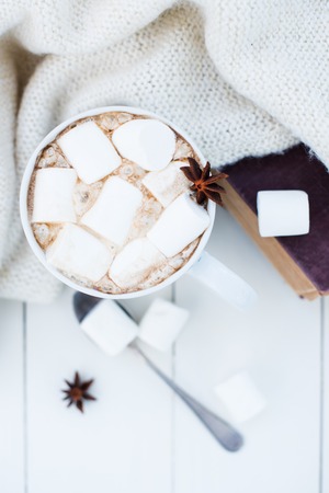 Cozy winter home background, cup of hot cocoa with marshmallow, old vintage books and warm knitted sweater on white painted wooden board background.の写真素材