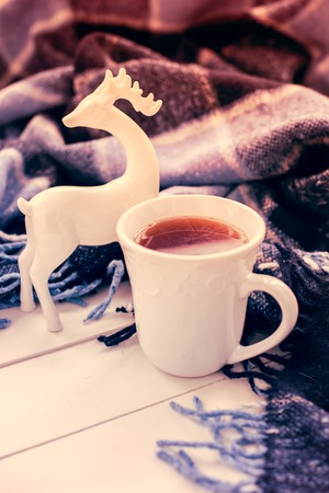 Beautiful white cup of hot tea, ceramic Christmas deer and a blue checkered woolen plaid. Cozy winter home arrangement hipster style.の写真素材