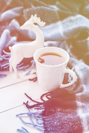 Beautiful white cup of hot tea, ceramic Christmas deer and a blue checkered woolen plaid. Cozy winter home arrangement hipster style.の写真素材