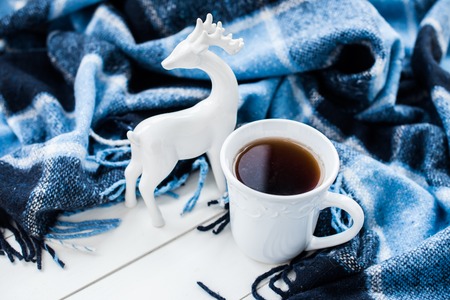 Beautiful white cup of hot tea, ceramic Christmas deer and a blue checkered woolen plaid. Cozy winter home arrangement.の写真素材