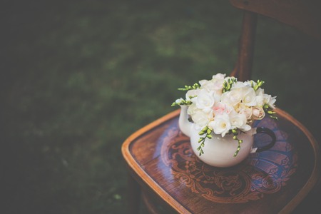 Summer wedding party festive decor, lovely fresh bouquet of white summer roses and freesias in vintage enamel tea pot on an old brown wooden chair. Retro style outdoor decoration, grass background.の写真素材