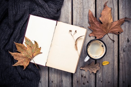 Open empty vintage book, knitted sweater with autumn leaves and coffee mug on old wooden board backgroundの写真素材