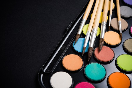 New watercolor paint set and brushes on artist's work desk, creative art tools isolated on black...