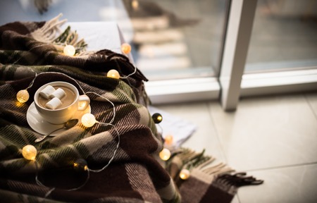 Cozy winter home, cup of coffee with marshmallows, warm blanket and Christmas lights