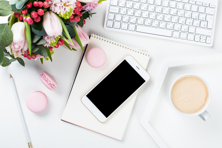 Lady bloggers work desk with pink flowers and macaron cakes on white table background, feminine home office workspce with coffee and smartphone mock-up