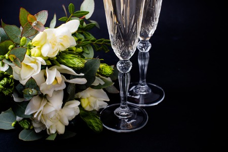 Two glasses of champagne and bouquet of white flowers on black background, sparkling wine and festive decoration with freesias and orchidsの写真素材