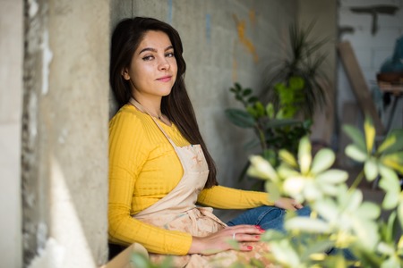 Young beautiful lady artist in apron with paint stains sitting on the floor in her studio