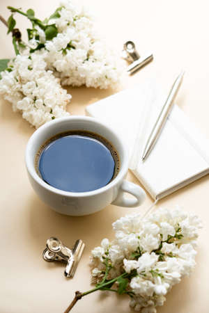 Beige and white work space with coffee cup, notepad and lilac flowersの写真素材