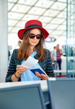 Beautiful young business woman in red hat in the airport at front desk service registers to airplane flight with passport and boarding pass documents in hands.の写真素材