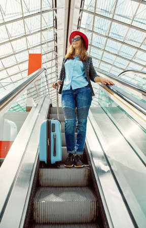 Beautiful young girl traveler in red hat with blue suitcase go down on escalator in the airport.の写真素材