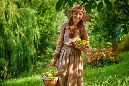 Pretty smiling teenager girl in the garden with basket with apples harvest picked from apple tree. Warm sunny day.の写真素材