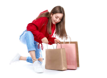 Young beautiful smiling girl in red sweetshot and blue jeans with paper bags for shopping sitting on the floor happy after buying. Shopping concept, isolated on white background.の写真素材