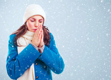 Frozen girl in blue coat, white hat and scarf heats hands among winter snowstorm.