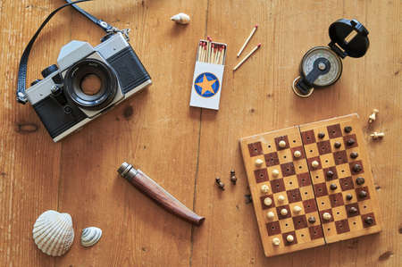 Top view of retro travel equipment on a wooden table. Vintage camera, matches, knife, mussel, chess board and compass on a wooden table.の写真素材