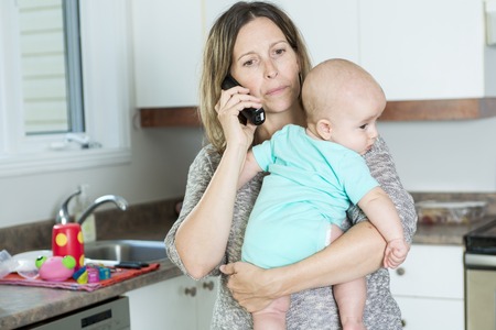 A woman on the phone while holding her baby in her arms in the kitchen