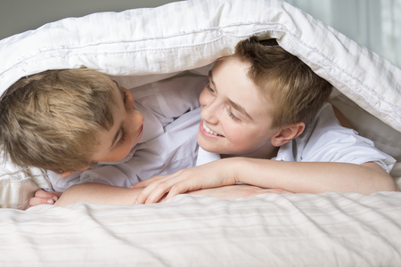 two boys hiding in bed under a white blanket or coverlet.の写真素材