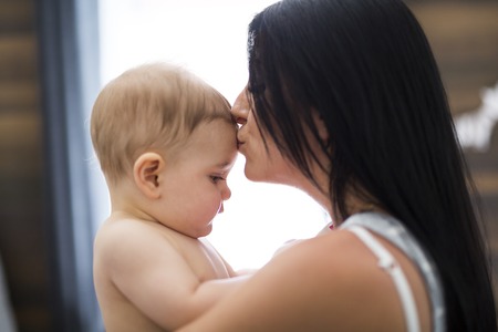 Toddler baby in a tender embrace of mother at the windowの写真素材