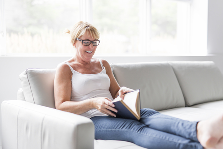 Shot of a mature woman reading her favorite novel while at home in living room.の写真素材